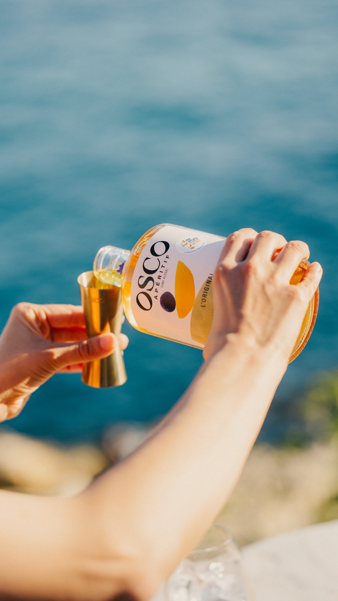 Main versant l'apéritif sans alcool OSCO Original dans un doseur doré, en extérieur avec vue sur la mer avec beaucoup de soleil